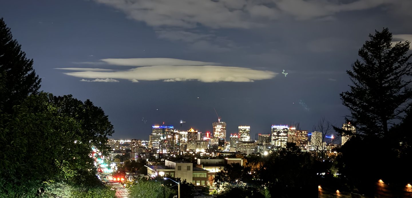 Downtown lit up at night under a long lenticular cloud, framed by dark trees